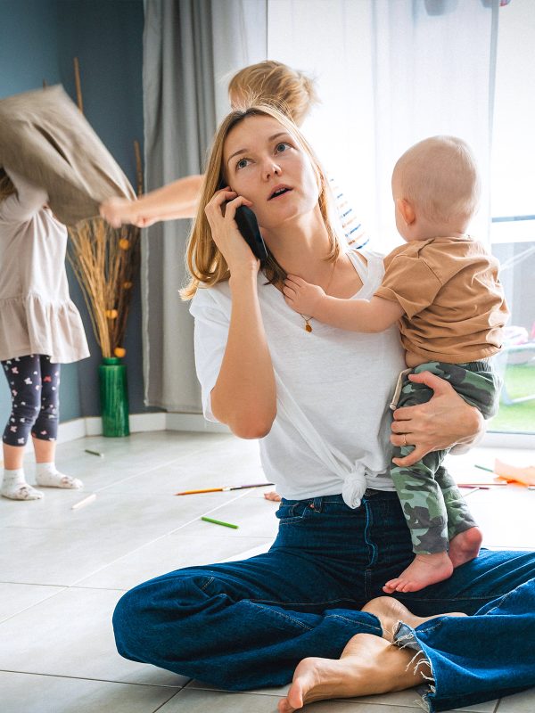 FAMILY COMMUNITY Young mum holding her infant with her young children having a pillow fight in the background while she is on the phone